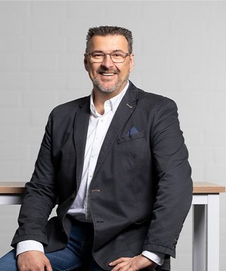 Der Mann trägt eine Brille und hat graue Haare, während er in einem modernen, hellen Raum freundlich in die Kamera schaut.