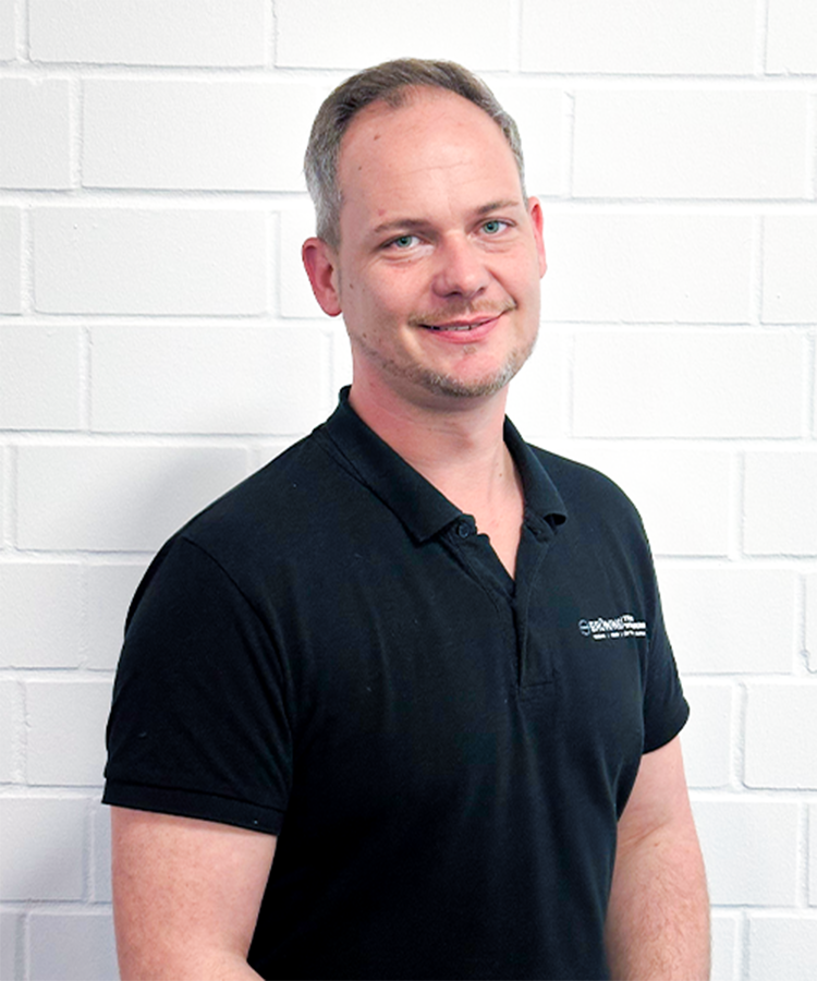 In einem schwarzen Poloshirt lächelt ein Mann mit grauen Haaren vor einer weißen Wand, die eine freundliche Atmosphäre schafft.