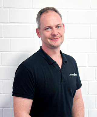 In einem schwarzen Poloshirt lächelt ein Mann mit grauen Haaren vor einer weißen Wand, die eine freundliche Atmosphäre schafft.