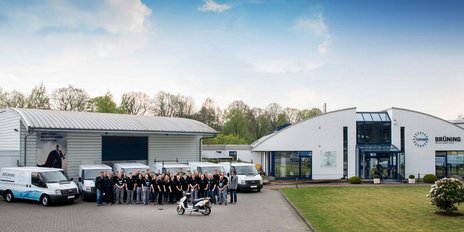 Das Team von Brüning posiert lächelnd vor ihrem Bürogebäude, umgeben von grüner Landschaft und Fahrzeugen.