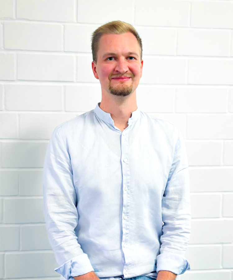 Ein junger Mann mit blonden Haaren und einem Bart lächelt freundlich vor einer weißen Ziegelwand.