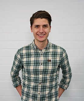 Ein junger Mann mit einem freundlichen Lächeln trägt ein kariertes Hemd und steht vor einer weißen Wand.