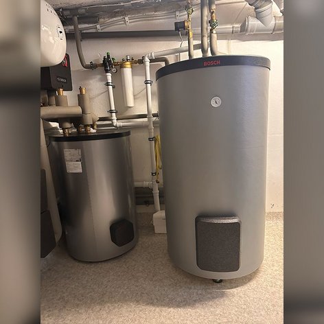 Ein heller Keller mit zwei Wasserboilern, die auf einem hellen Boden stehen, umgeben von verschiedenen Rohrleitungen und Ventilen.