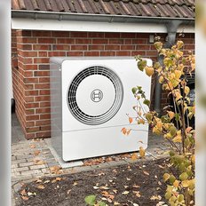 Die weiße Wärmepumpe ist an einer roten Ziegelwand installiert, während gelbe Blätter im Vordergrund fallen.