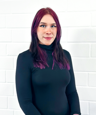 Eine Person mit langen, lila gefärbten Haaren steht vor einer weißen Wand und trägt ein schwarzes Oberteil.