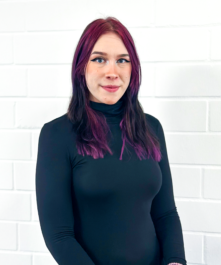 Eine Person mit langen, lila gefärbten Haaren steht vor einer weißen Wand und trägt ein schwarzes Oberteil.