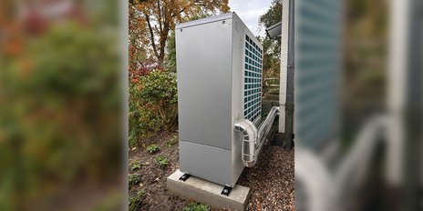 Im Hintergrund sind herbstliche Bäume zu sehen, während die Wärmepumpe in einem gepflegten Garten platziert ist.