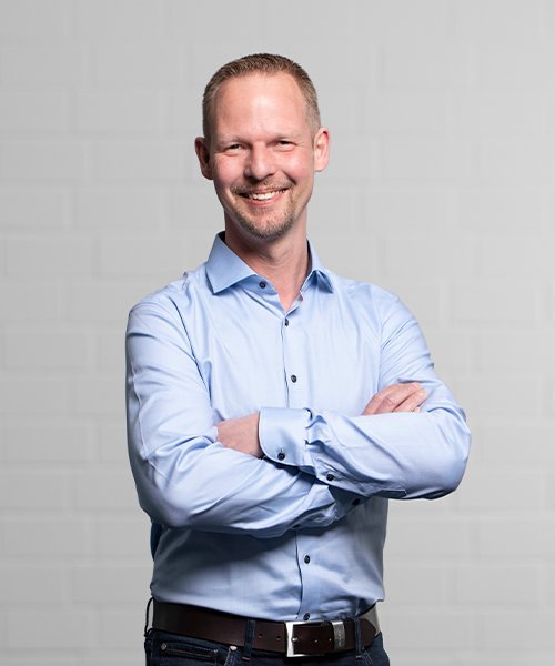 Der Mann in einem hellblauen Hemd zeigt ein freundliches Lächeln, während er entspannt vor einer neutralen Hintergrundwand posiert.