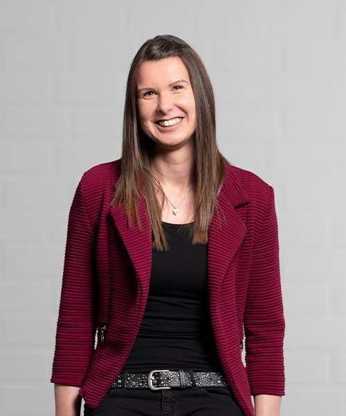 Mit einem freundlichen Lächeln präsentiert sich die Frau in einer modischen roten Jacke vor einer hellen, minimalistischen Wand.