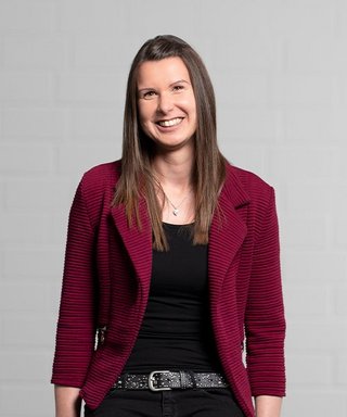 Mit einem freundlichen Lächeln präsentiert sich die Frau in einer modischen roten Jacke vor einer hellen, minimalistischen Wand.