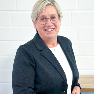 Die Frau mit blonden Haaren und Brille zeigt ein freundliches Lächeln, während sie an einer hellen Wand steht.