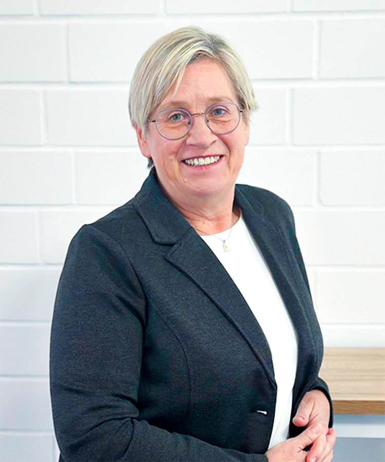 Die Frau mit blonden Haaren und Brille zeigt ein freundliches Lächeln, während sie an einer hellen Wand steht.