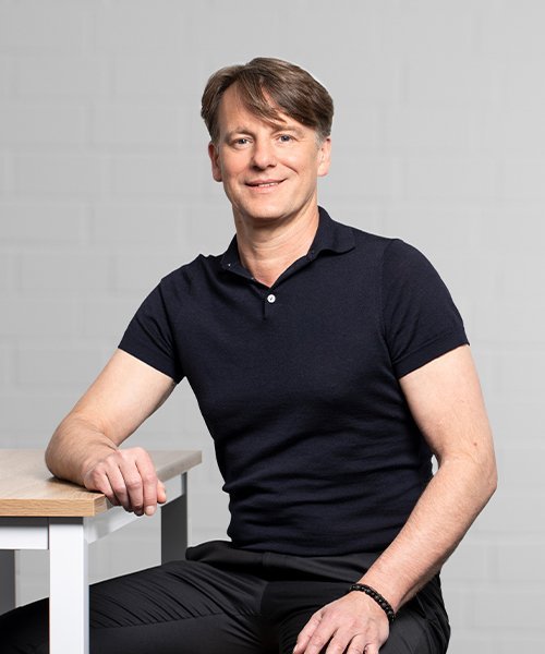 Ein Mann mit kurzem, braunem Haar sitzt entspannt an einem Tisch und lächelt freundlich in die Kamera.