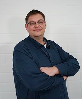 Der Mann trägt ein dunkelblaues Hemd und hat einen freundlichen Gesichtsausdruck, während er vor einer hellen Wand posiert.