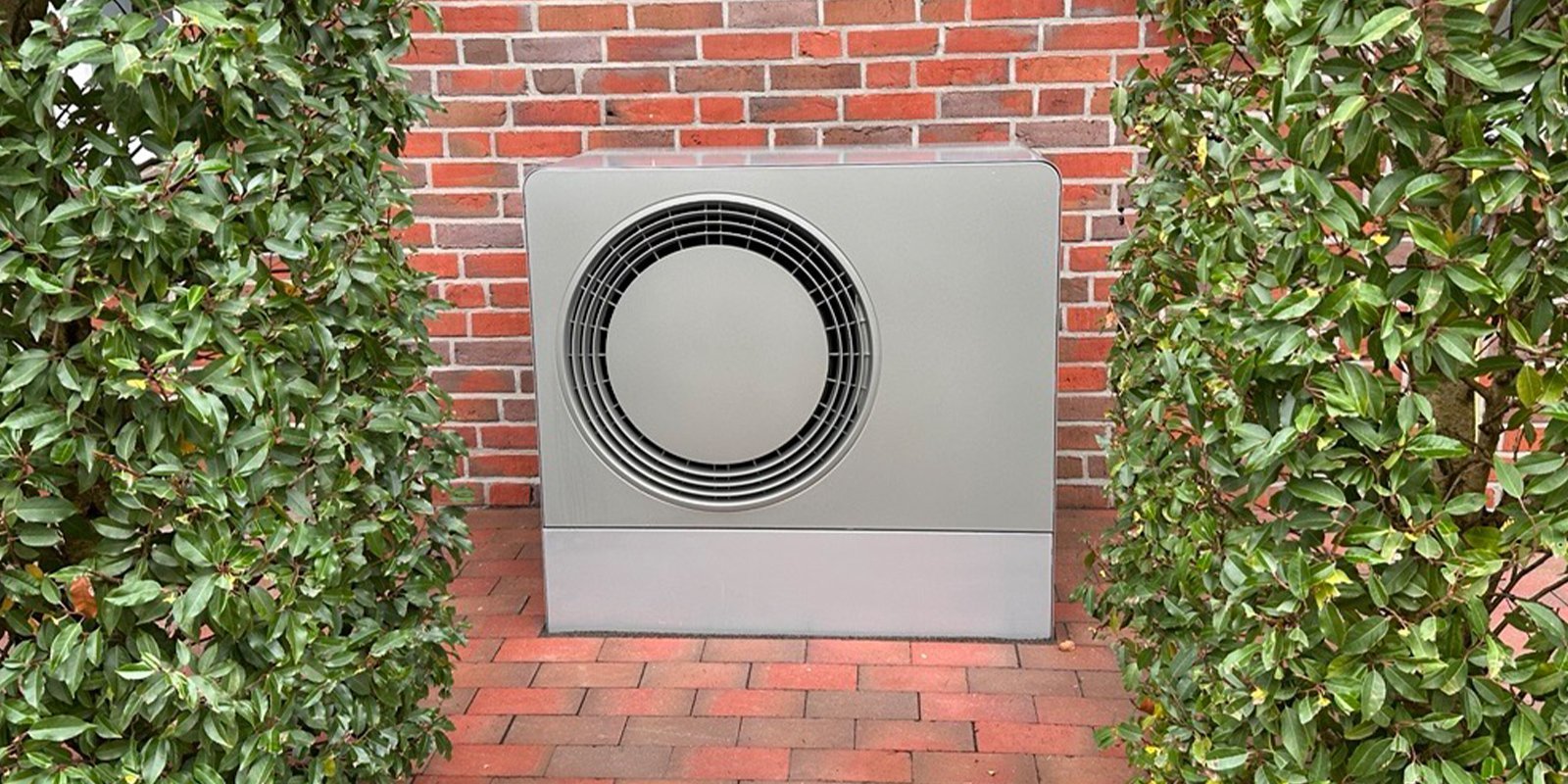 Die Klimaanlage ist gut sichtbar, umgeben von üppigem Grün und einer rustikalen Backsteinmauer im Hintergrund.