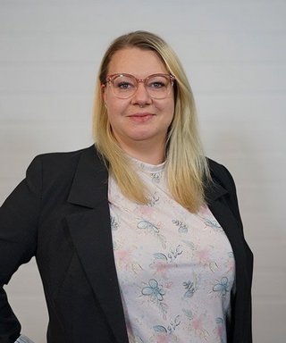 Eine Frau mit blonden Haaren trägt eine Bluse mit Blumenmuster und einen schwarzen Blazer, lächelt freundlich in die Kamera.