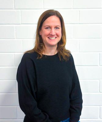 Eine lächelnde Frau mit langen, braunen Haaren trägt einen schwarzen Pullover und steht vor einer weißen Ziegelwand.