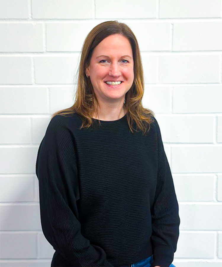 Eine lächelnde Frau mit langen, braunen Haaren trägt einen schwarzen Pullover und steht vor einer weißen Ziegelwand.