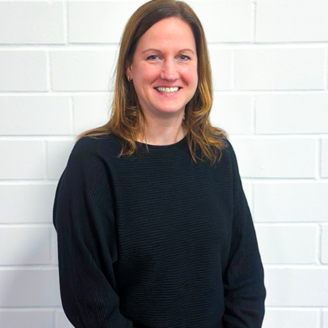 Eine lächelnde Frau mit langen, braunen Haaren trägt einen schwarzen Pullover und steht vor einer weißen Ziegelwand.