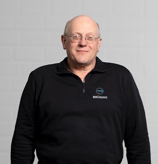 Porträt eines Mannes mit Brille und freundlichem Ausdruck Der Mann mit Glatze und Brille lächelt leicht, während er in einem schwarzen Oberteil vor einem neutralen Hintergrund posiert.