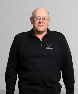 Der Mann mit Glatze und Brille lächelt leicht, während er in einem schwarzen Oberteil vor einem neutralen Hintergrund posiert.