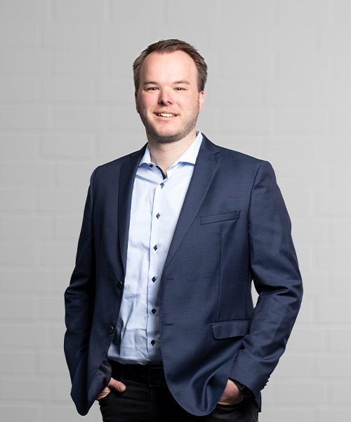 Der Mann trägt ein hellblaues Hemd unter einem dunklen Blazer und steht entspannt mit einer Hand in der Hosentasche.