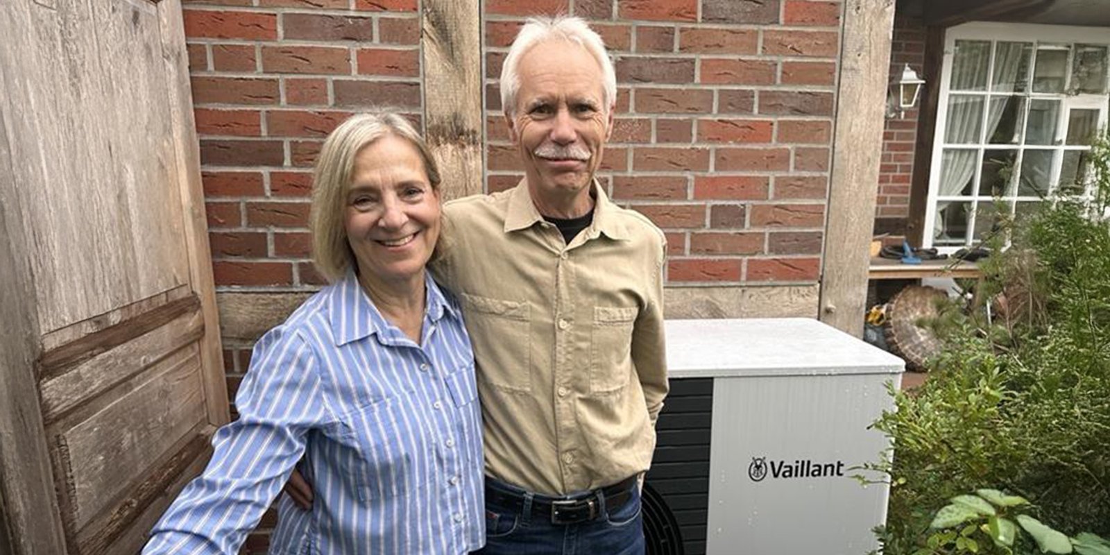 In einem Garten stehen ein Mann und eine Frau, beide lächeln, während sie neben einem Heizgerät von Vaillant stehen.