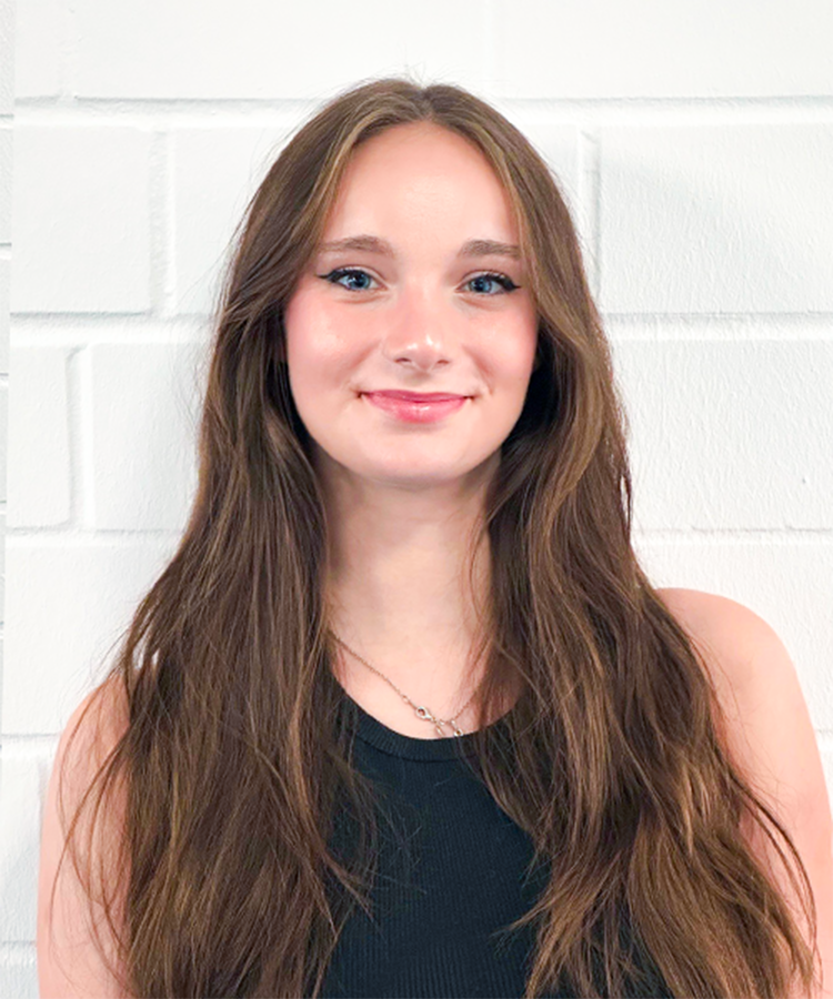 Eine junge Frau mit langen, braunen Haaren lächelt freundlich vor einer weißen Backsteinwand.