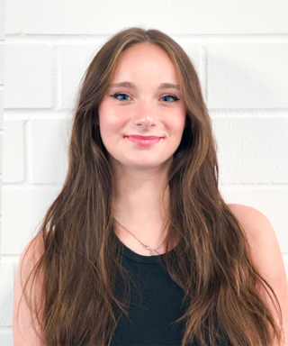 Eine junge Frau mit langen, braunen Haaren lächelt freundlich vor einer weißen Backsteinwand.