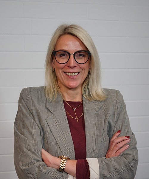 Eine blonde Frau mit Brille lächelt selbstbewusst, die Arme vor der Brust verschränkt, vor einer weißen Wand.