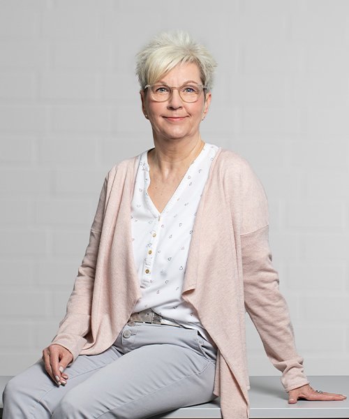 Eine Frau mit blonden, kurzen Haaren und Brille sitzt entspannt auf einem Tisch, lächelnd und in einem hellen Outfit.