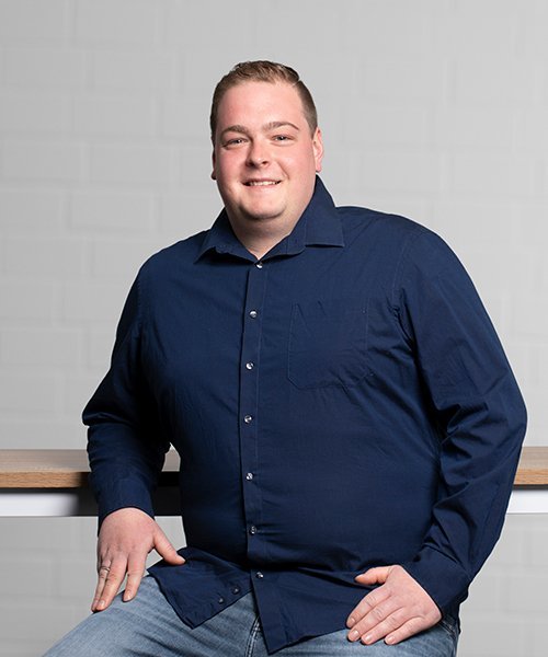 Ein lächelnder Mann sitzt entspannt an einem Tisch, trägt ein dunkelblaues Hemd und hat eine freundliche Ausstrahlung.