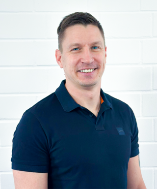 Ein Mann mit kurzem, braunem Haar lächelt freundlich vor einer weißen Ziegelwand, gekleidet in ein schwarzes Polohemd.