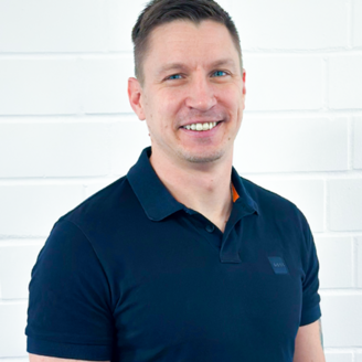 Ein Mann mit kurzem, braunem Haar lächelt freundlich vor einer weißen Ziegelwand, gekleidet in ein schwarzes Polohemd.