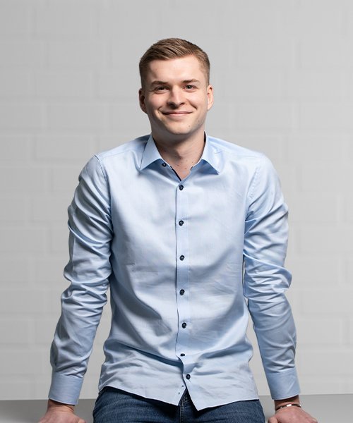Ein lächelnder junger Mann in einem hellblauen Hemd posiert an einem Tisch, umgeben von einer neutralen Wand.