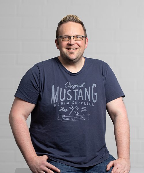 Ein lächelnder Mann mit Brille trägt ein dunkelblaues T-Shirt mit dem Aufdruck 'Mustang Denim Supplies'.
