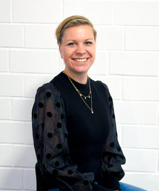 Eine lächelnde Frau mit blonden Haaren sitzt vor einer weißen Ziegelwand, gekleidet in einem schwarzen Pullover mit gepunkteten Ärmeln.