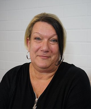 Die Frau hat eine entspannte Ausstrahlung und trägt ein schwarzes Oberteil, während sie direkt in die Kamera blickt.