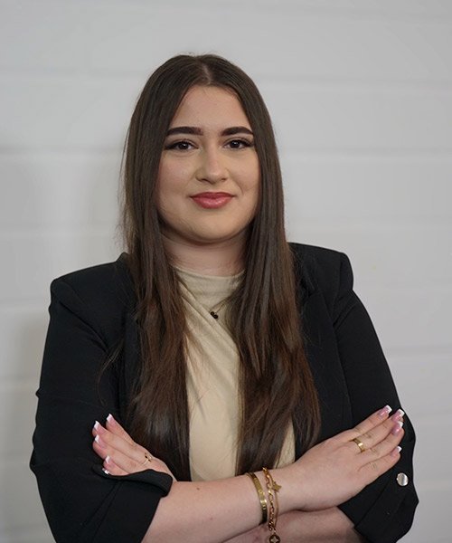 Die Frau trägt einen schwarzen Blazer und ein beiges Oberteil, während sie in die Kamera schaut und eine positive Ausstrahlung hat.