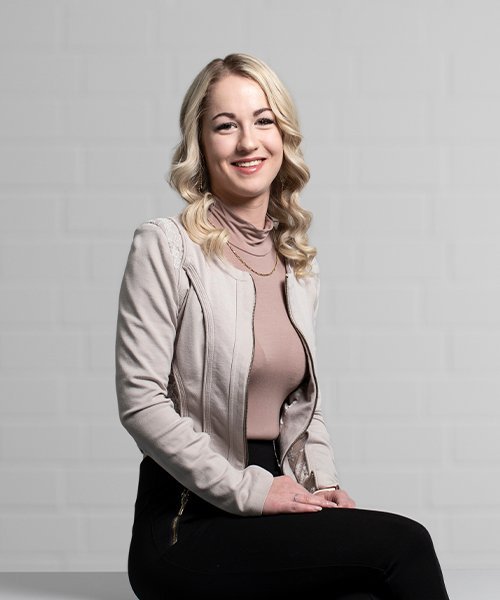 Die Frau trägt eine elegante, helle Jacke und posiert selbstbewusst, während sie in die Kamera lächelt.