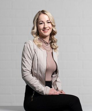 Die Frau trägt eine elegante, helle Jacke und posiert selbstbewusst, während sie in die Kamera lächelt.