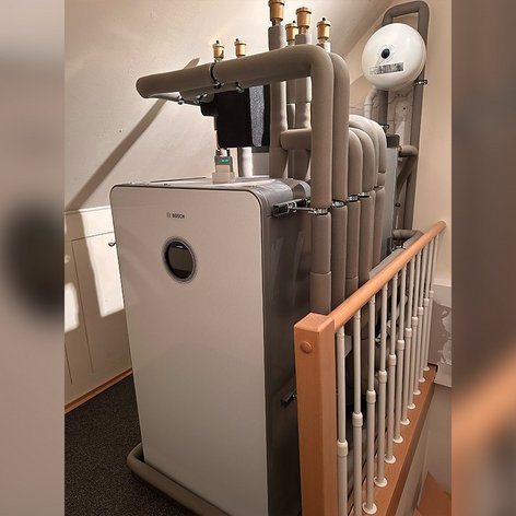 Die Heizungsanlage ist in einem Wohnraum installiert, mit einem Holzgeländer und Rohren, die die technische Ausstattung zeigen.