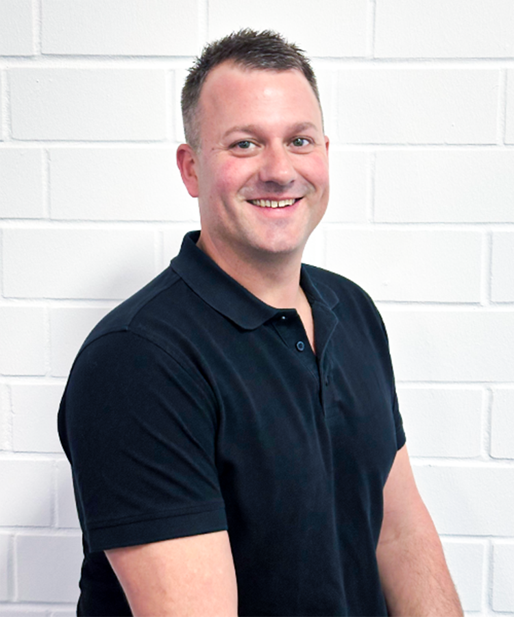 Der Mann mit kurzen Haaren und einem breiten Lächeln posiert entspannt vor einer hellen, strukturierten Wand.